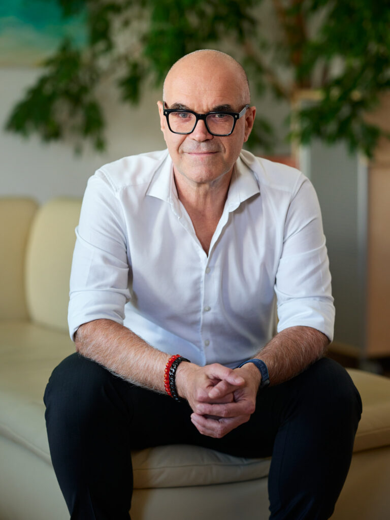 Günter Neubauer bietet Systemisches Coaching für Führungskräfte an: Porträtfoto, aufrecht sitzend, konzentrierter Blick in die Kamera, dunkles Hemd, im Hintergrund unscharfe Bücherregale.