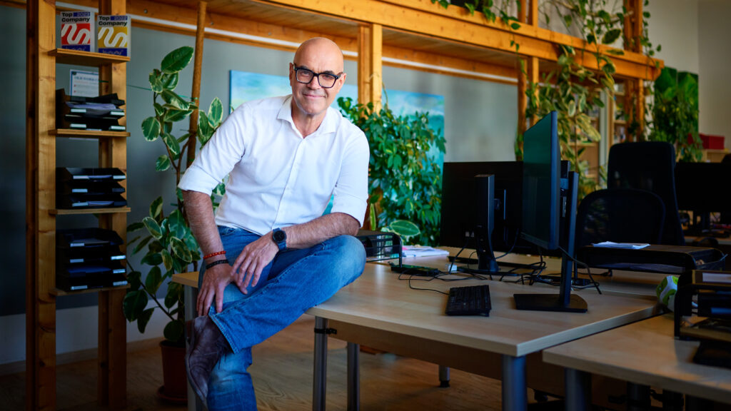 Günter Neubauer, Sparringspartner für Führungskräfte, sitzt entspannt lächelnd in seinem Büro zwischen Holzbalken und Pflanzen, im Hintergrund Bildschirme und ein Bücherregal.